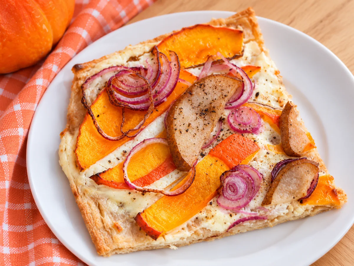 Ein Stück Kürbis-Flammkuchen mit gebackenen Kürbisspalten, Birne und roten Zwiebelringen auf einem weißen Teller neben einem orange karierten Tuch.