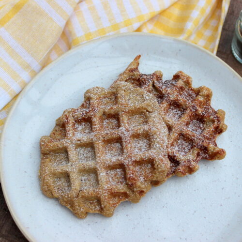 Zwei Vanille-Joghurt-Waffeln auf einem beigen Steingut-Teller auf einem Holztisch mit einem gelb-weiß karierten Geschirrtuch und einem kleinen Gläschen Vanillepulver daneben.